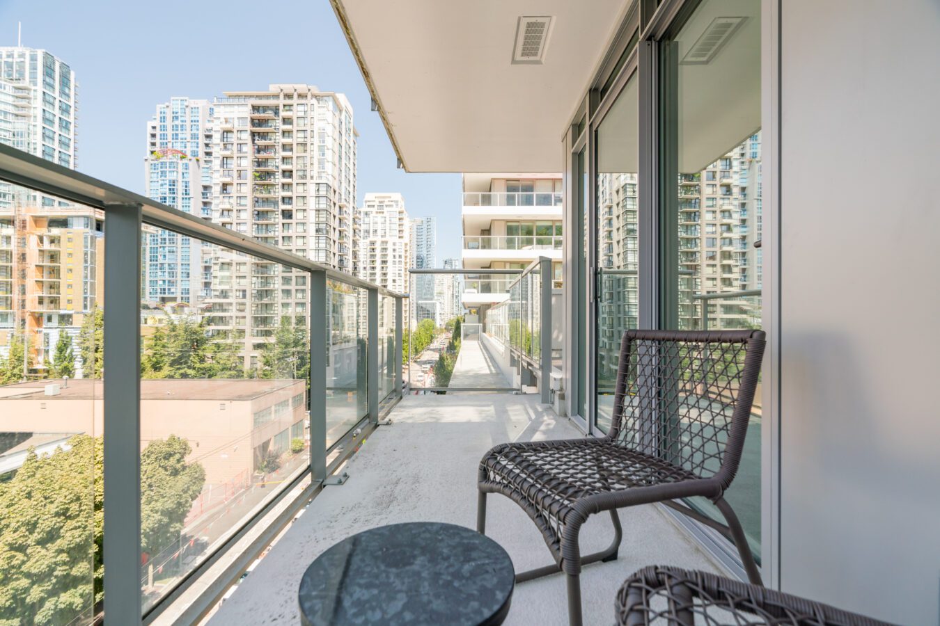 A modern balcony with glass railings and two wicker chairs facing a small round table, overlooking a cityscape with tall apartment buildings and trees.