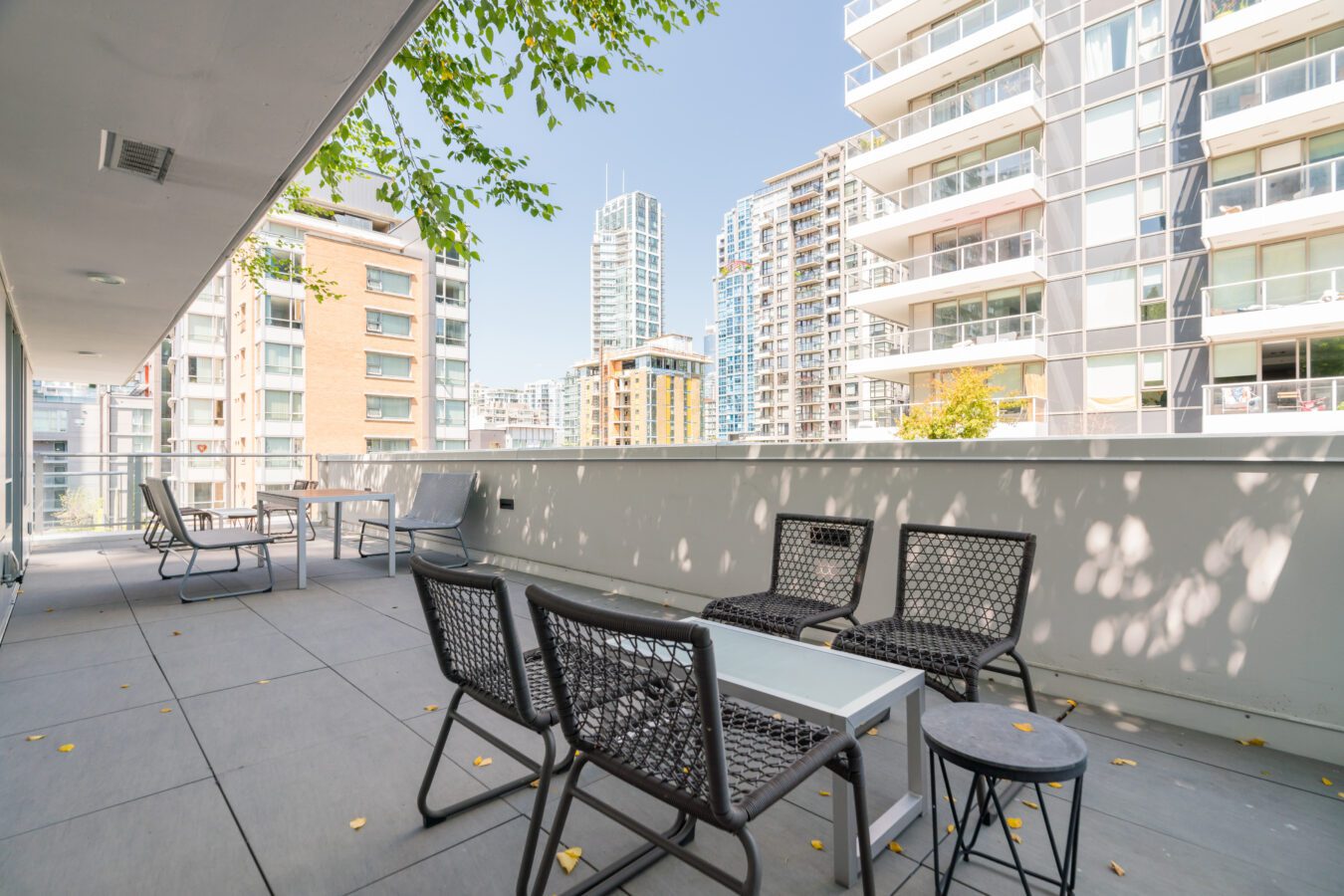 Spacious apartment balcony with modern black chairs and a white table, overlooking tall residential buildings under a clear blue sky. Some tree branches and scattered yellow leaves are visible.