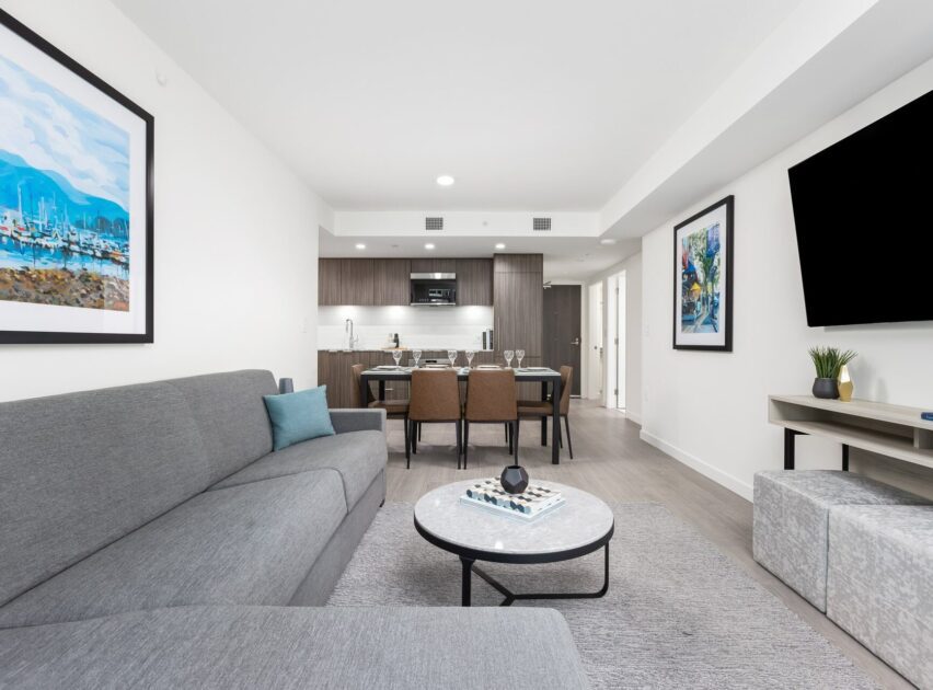 Modern living room with a gray sofa, round coffee table, wall-mounted TV, and artwork. In the background, there is a dining table and a kitchen with dark cabinets and stainless steel appliances.