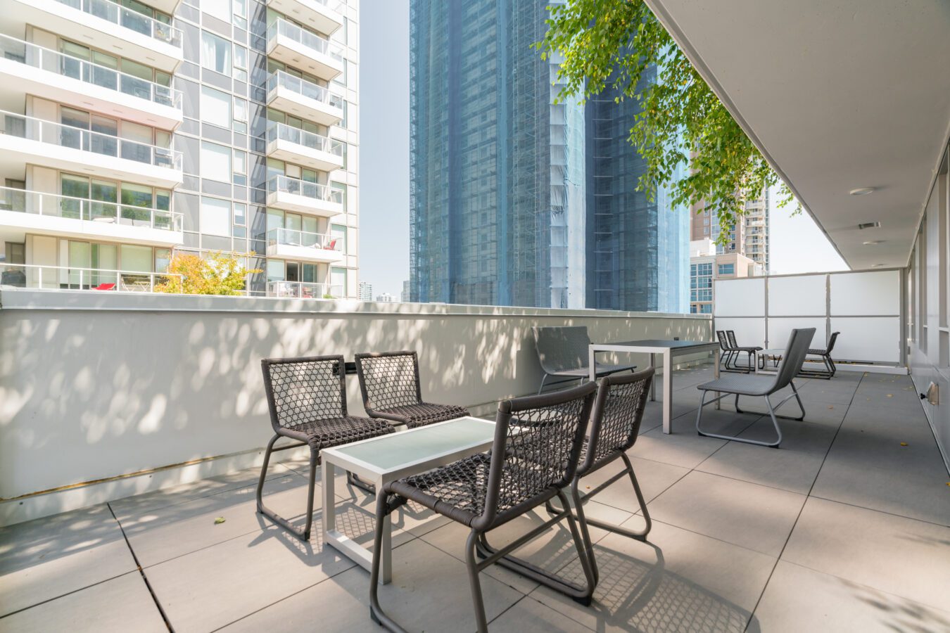 Outdoor terrace with modern black chairs and white tables, surrounded by high-rise buildings. The area is shaded partially by trees, with sunlight casting shadows on the tiled floor.