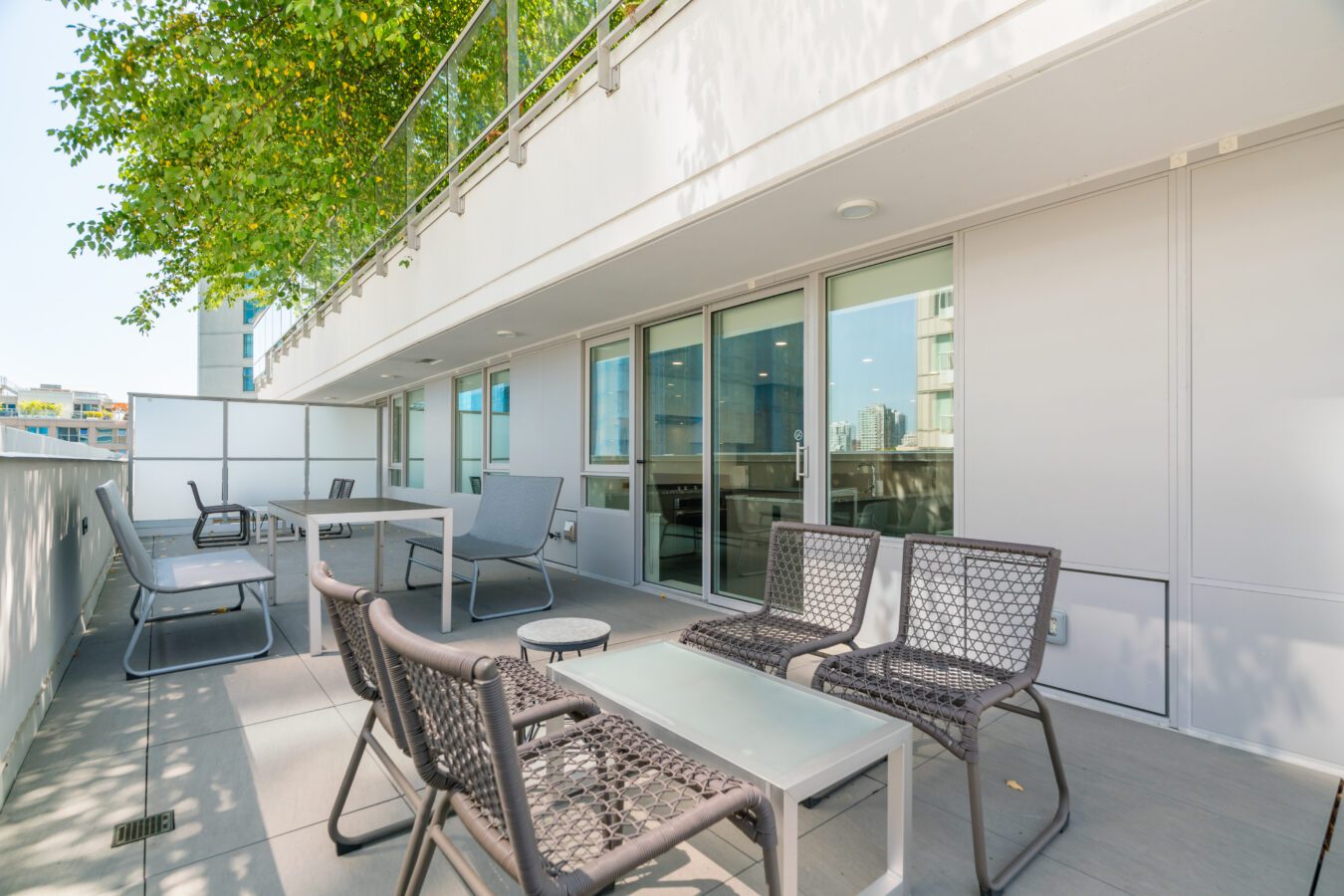 Modern apartment balcony with gray chairs, tables, and glass doors, partially shaded by a tree. The bright, spacious outdoor area overlooks nearby buildings and features minimalist outdoor furniture.