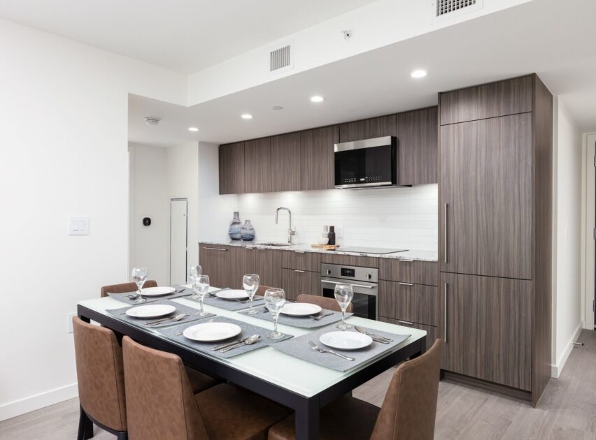 Modern kitchen and dining area with a set table for six, featuring brown chairs, a gray table runner, wood cabinetry, stainless steel appliances, and white tile backsplash under recessed ceiling lights.