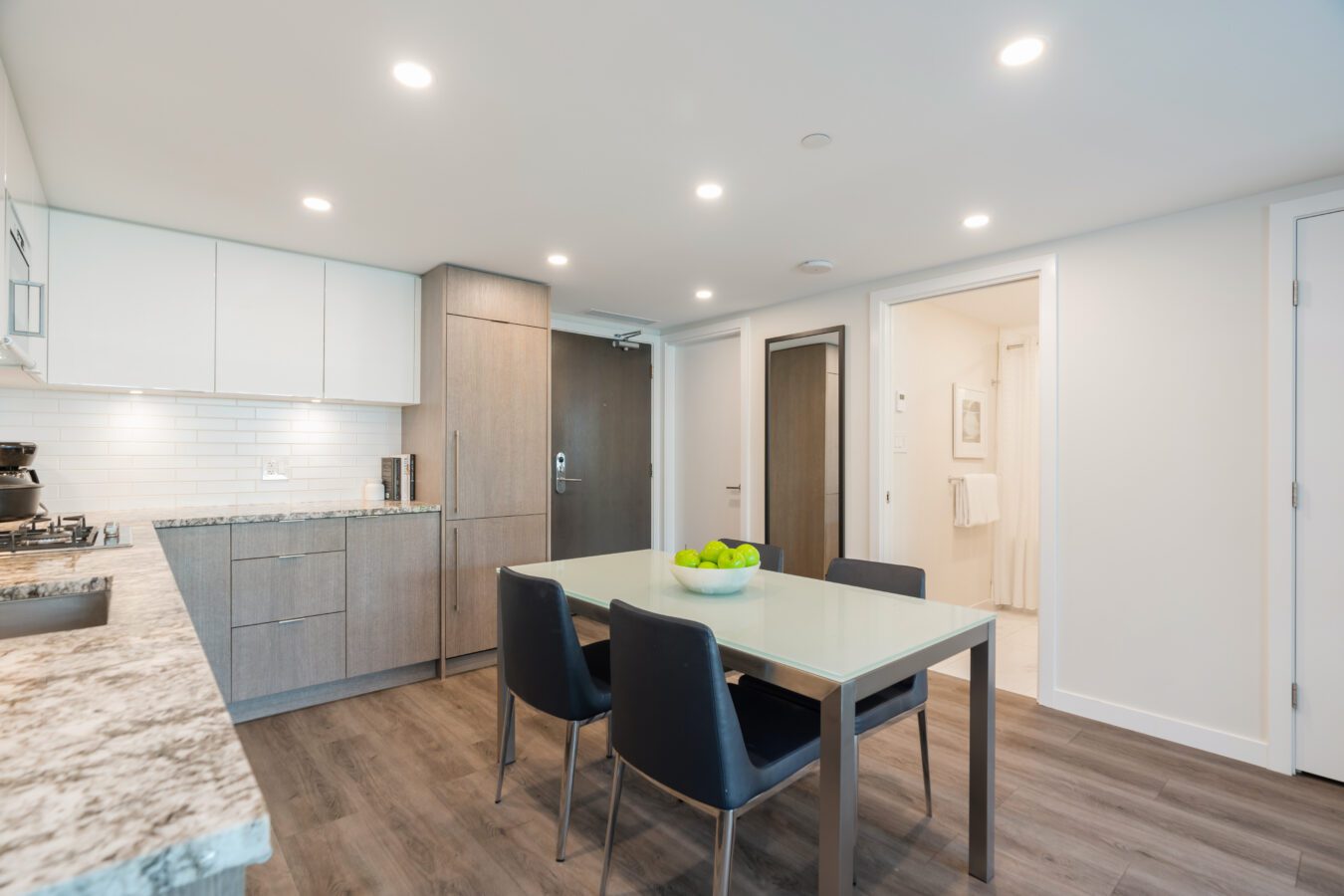 Modern kitchen and dining area with wood floors, light wood cabinets, a marble countertop, and a glass table with four black chairs. A bowl of green apples sits at the center of the table. Doorway leads to a bathroom.