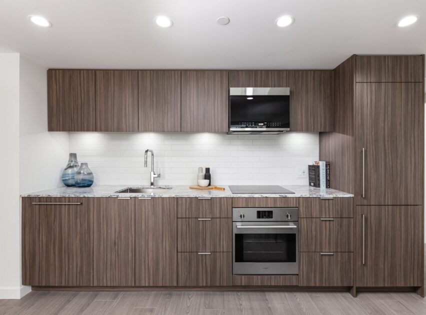 Modern kitchen with wood-paneled cabinets, built-in stainless steel oven, and microwave. White tile backsplash, marble countertop, sink, and minimal decor including vases and cutting boards. Bright overhead lighting.