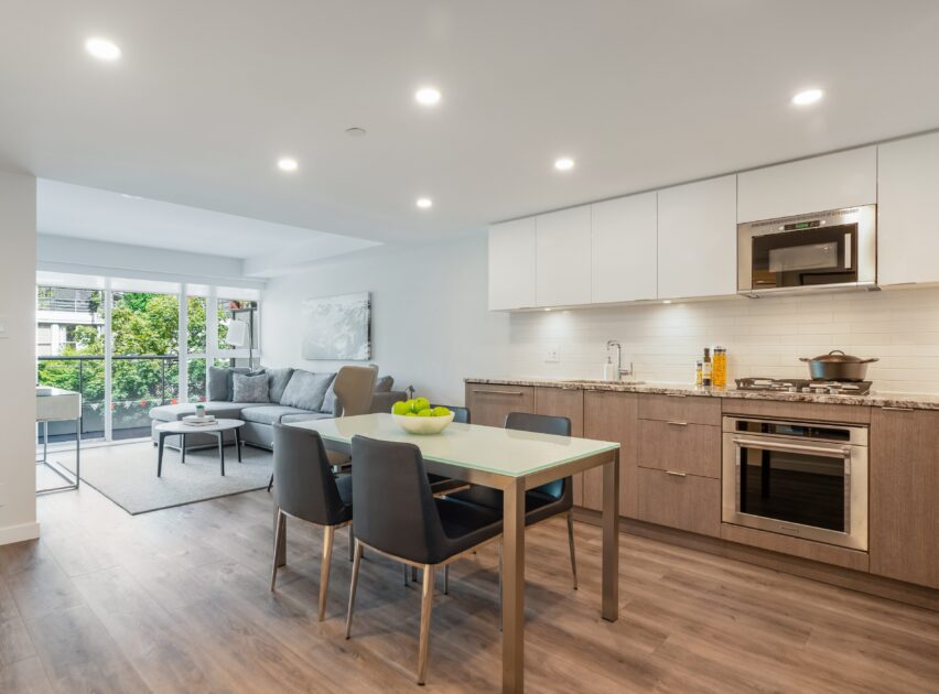 Modern open-concept kitchen and living room with wood flooring, sleek cabinets, a dining table with four chairs, a gray sofa, and large windows letting in natural light.