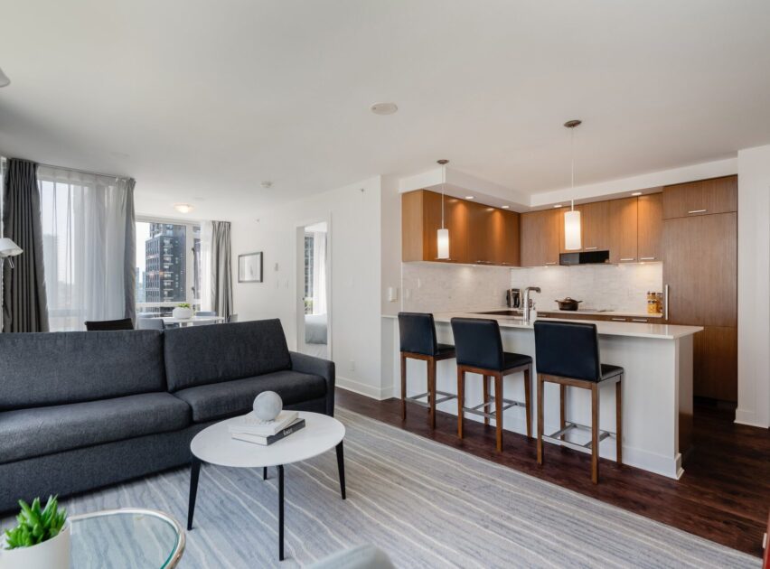 Modern open-concept apartment with a gray sofa, round coffee table, and kitchen area featuring wood cabinets, white island with three black barstools, pendant lights, and large windows letting in natural light.
