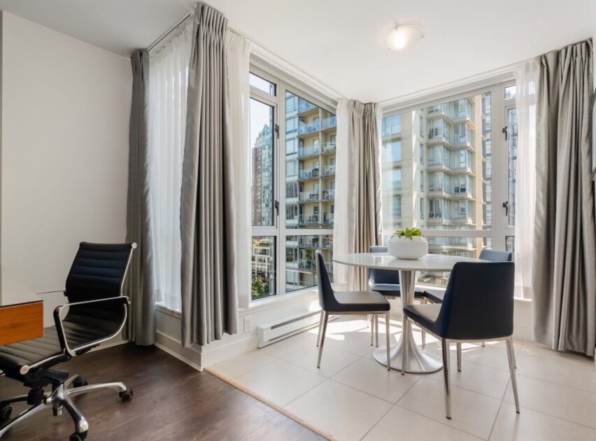 Modern dining area with a round table, three black chairs, a potted plant, large corner windows with city views, light tile floor, and an office chair on dark wood flooring in the corner.