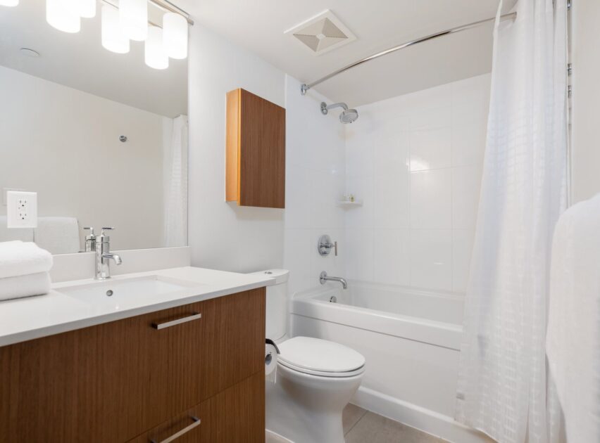 A modern bathroom with a white bathtub and shower, a white toilet, a wooden vanity with a white countertop, a wooden wall cabinet, and a large mirror under bright overhead lights.