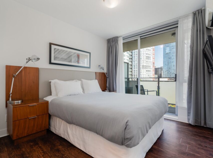 Modern bedroom with a large bed, gray bedding, two wooden nightstands with lamps, wall art above the bed, floor-to-ceiling window, balcony view of city buildings, dark wood floor, and wall-mounted TV.