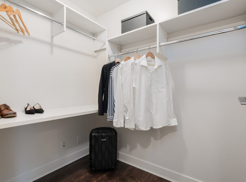 A spacious, modern walk-in closet with white shelves, a suitcase, a few shirts and sweaters on wooden hangers, two pairs of shoes on a shelf, and two black storage boxes on the upper shelf.