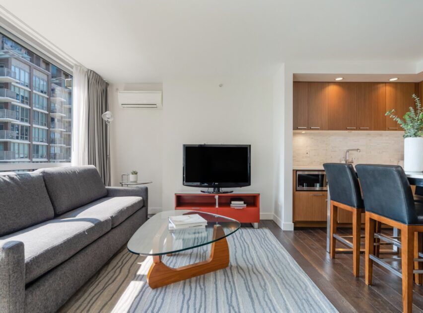 A modern living room with a gray sofa, glass coffee table, TV on a red stand, and a small kitchen area with wooden cabinets. Large windows let in bright natural light and show city buildings outside.