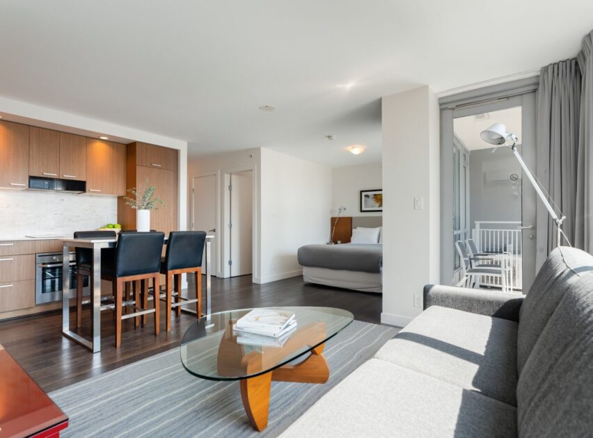 Modern open-plan apartment with a kitchen, dining table, and living area featuring a gray sofa and glass coffee table. In the background, a bedroom with a bed and a balcony with chairs are visible. Natural light fills the space.