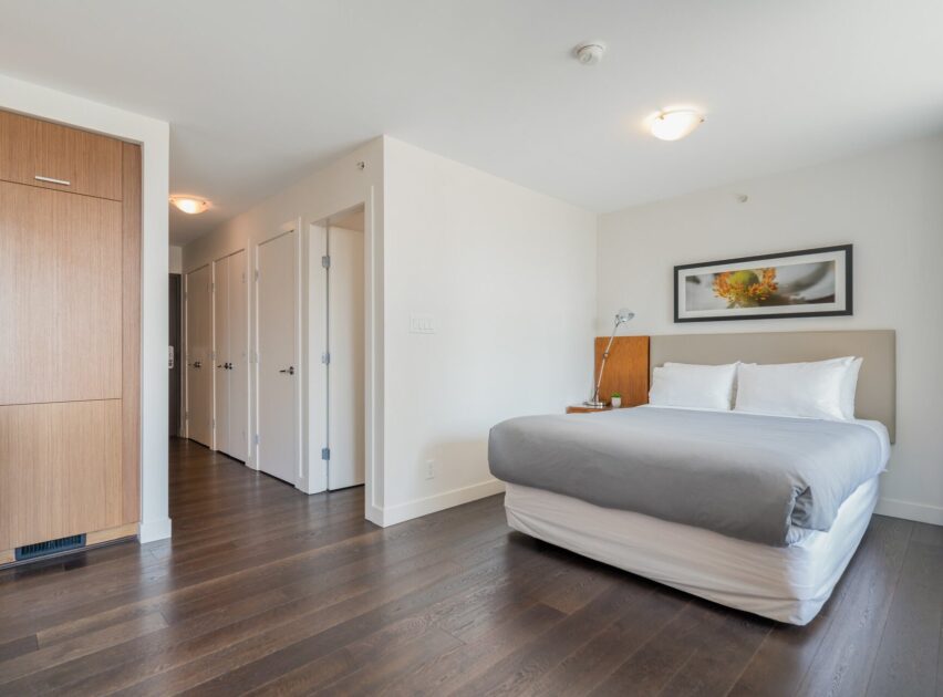 Minimalist bedroom with a neatly made bed, white walls, dark wood floor, a framed photo above the bed, and closed doors along one wall. A built-in wooden closet is visible on the left side.