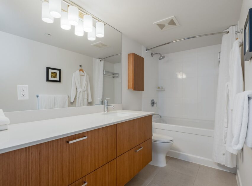 Modern bathroom with wooden cabinets, a large mirror, white countertop, towel rack, wall-mounted robe, bathtub with shower curtain, and bright overhead lighting. Walls and floor are light-colored, creating a clean look.