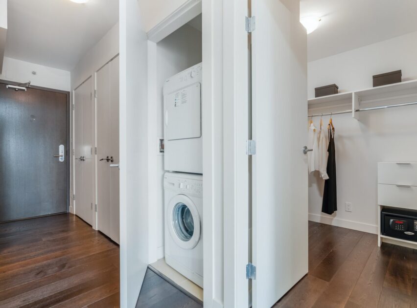 A modern apartment interior with dark wood floors, featuring a stacked washer and dryer in a closet, an open walk-in closet with hanging clothes, and a view of the entry door.