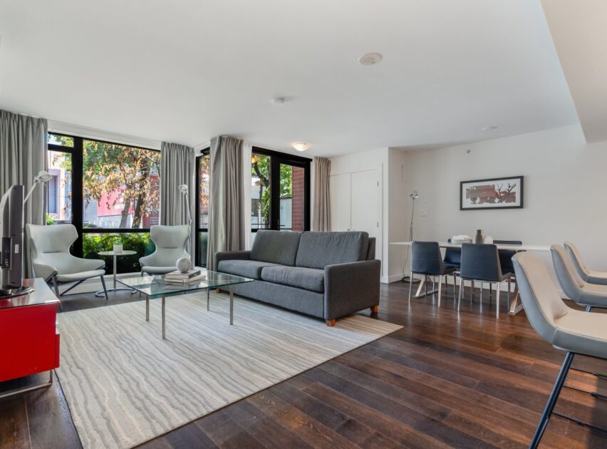 Modern living and dining area with large windows, a gray sofa, glass coffee table, flat-screen TV, dining table with six chairs, dark wood floors, and neutral décor. Natural light fills the space.