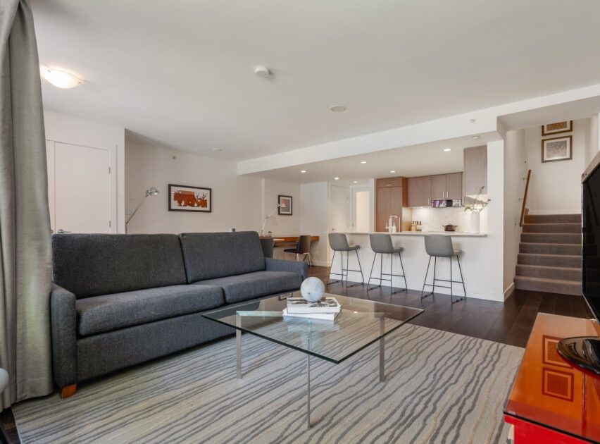 Modern open-concept living room with a gray sofa, glass coffee table, striped rug, and bar stools at a kitchen counter; kitchen and stairs are visible in the background.