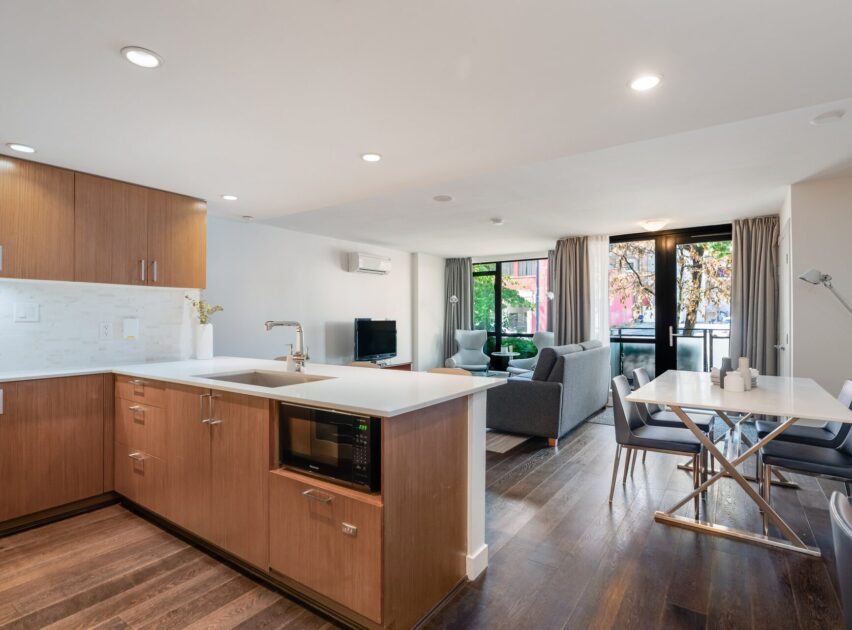 Modern open-concept kitchen and living area with wood cabinets, white countertops, dark wood flooring, a dining table, gray sofa, TV, and large windows showing greenery outside.
