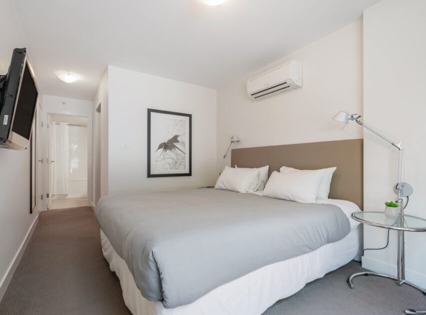 Minimalist bedroom with a large bed, gray bedding, two white pillows, wall-mounted lamps, a round side table with a small plant, wall art, air conditioner, and a TV mounted on the wall. White walls and neutral decor.