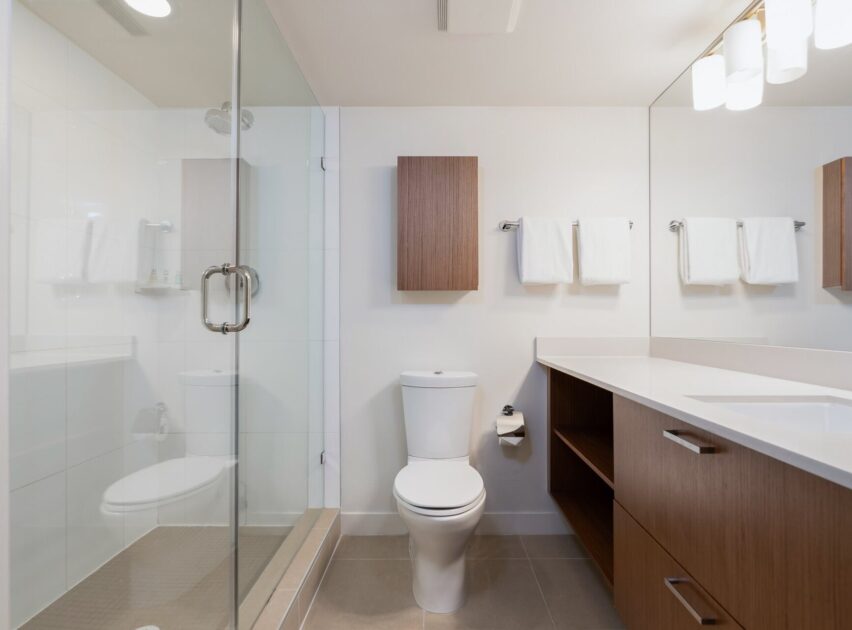Modern bathroom with a glass-enclosed shower, white toilet, wooden cabinets, three white towels on towel racks, and a wide countertop with built-in sink under a wall-mounted light fixture.