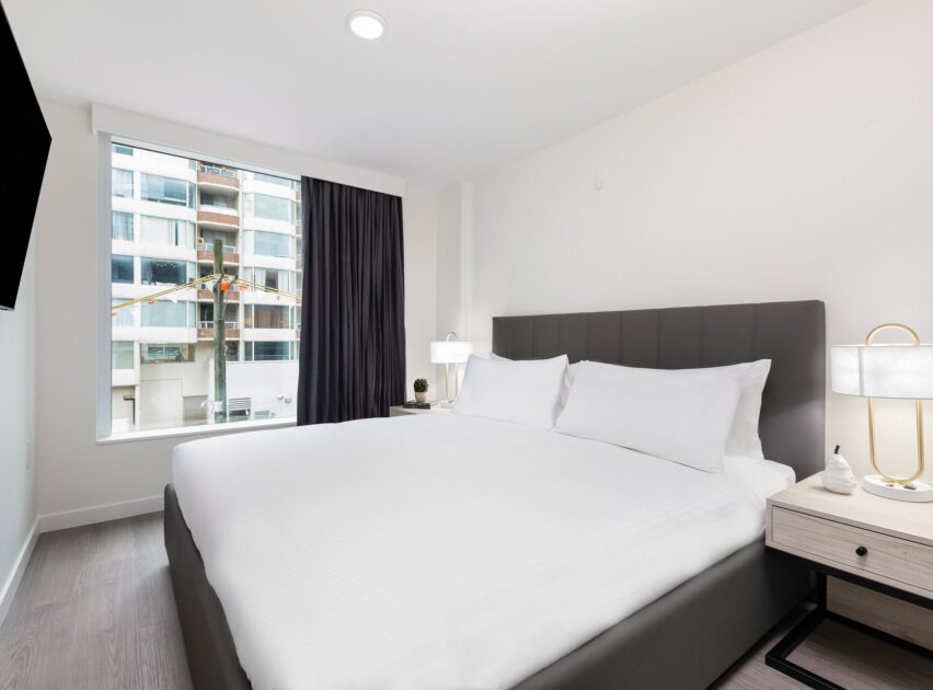 A modern hotel room with a large bed, white bedding, two bedside tables with lamps, a window with a city view, and a wall-mounted TV. The decor is minimalist with neutral colors.