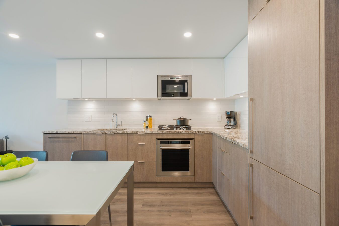 Modern kitchen with light wood cabinets, white countertops, built-in oven, microwave, and a coffee maker. A glass dining table with green apples in a bowl is in the foreground. Recessed lighting brightens the space.