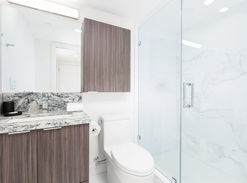Modern bathroom with a glass shower, white toilet, and a vanity with wood cabinets and a marble countertop. The space is bright with white walls and clean, minimalist design.