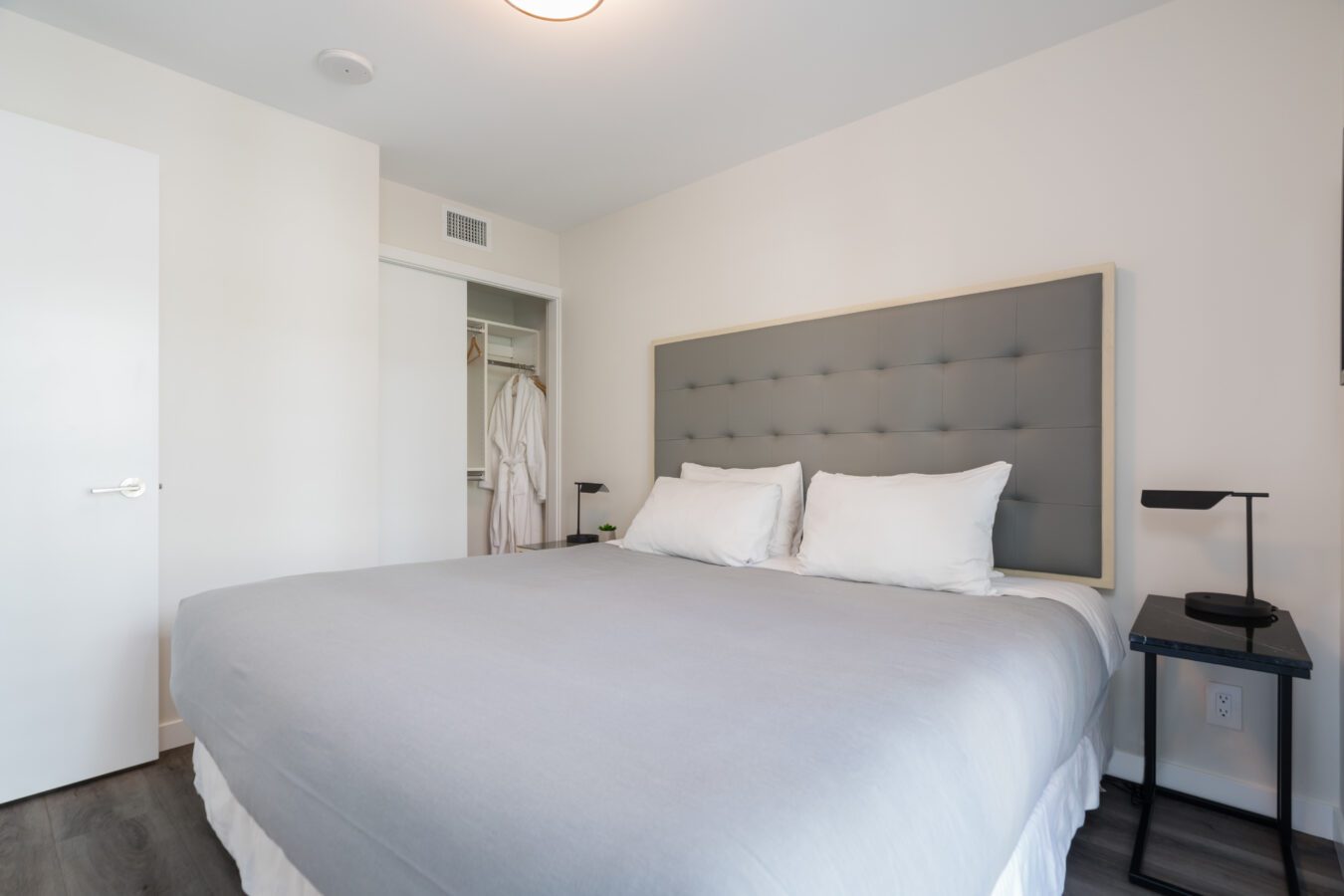 A modern bedroom with a large bed featuring a gray upholstered headboard, white bedding, and two black bedside tables with lamps. An open closet with white robes and shelves is visible in the background.