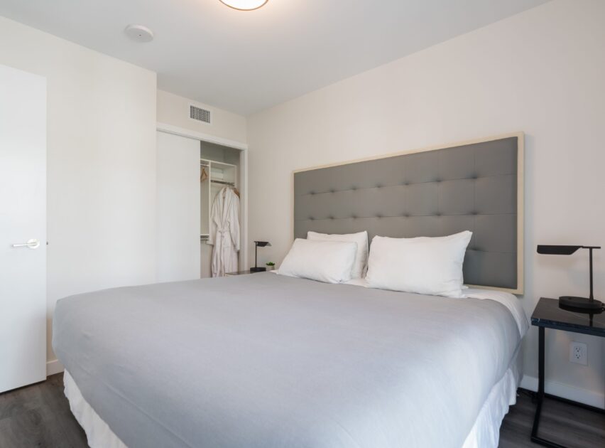 A tidy bedroom with a large bed featuring a gray tufted headboard, white and gray bedding, two black nightstands with lamps, an open closet with robes, and a white door. The room has white walls and a modern feel.