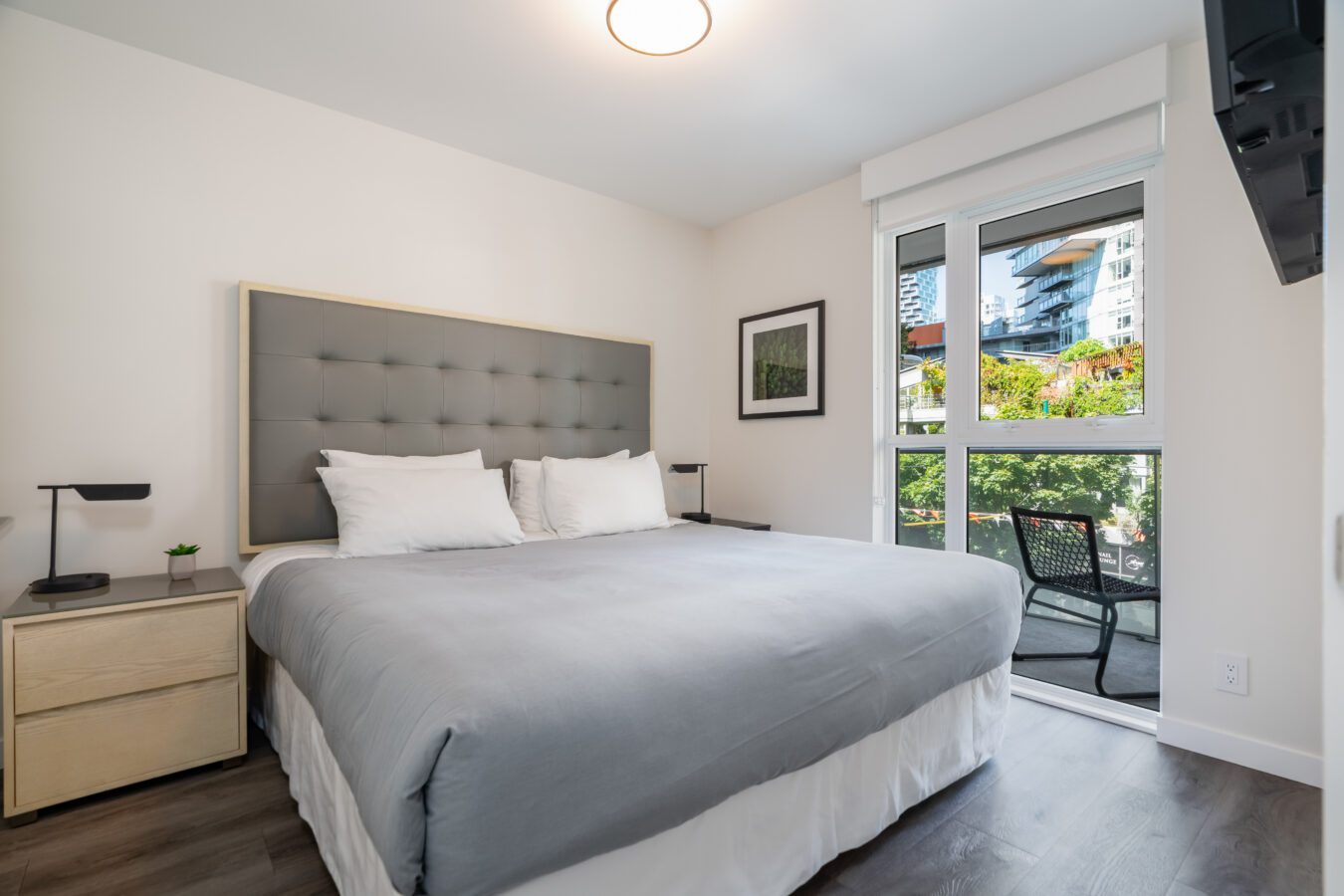 A modern bedroom with a large bed, gray tufted headboard, two bedside tables with lamps, a window with city and greenery views, and a chair on a small balcony. The room has white walls and wood flooring.