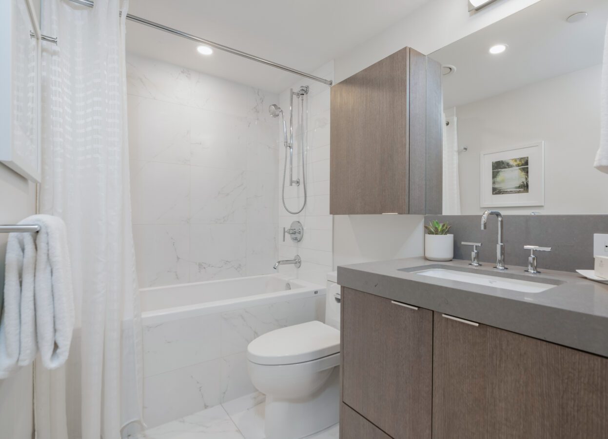 Modern bathroom with a gray vanity, white countertop, sink, wall-mounted faucet, toilet, and a bathtub-shower combo with white marble tile. A white shower curtain, towels, and a potted plant are also visible.
