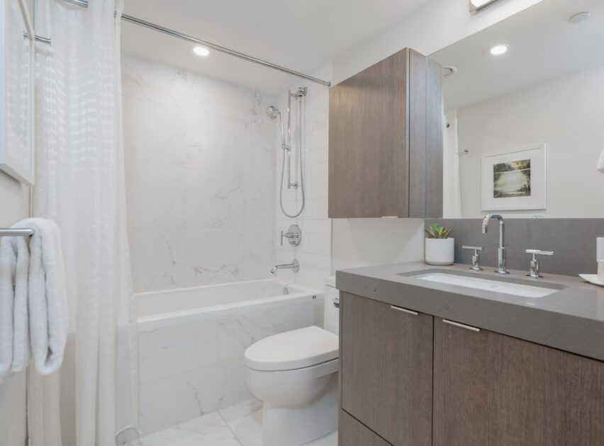 Modern bathroom with a bathtub and shower, gray vanity with sink, large mirror, white towels hanging on a rack, and a potted plant on the counter. Walls and floor are white with marble pattern.