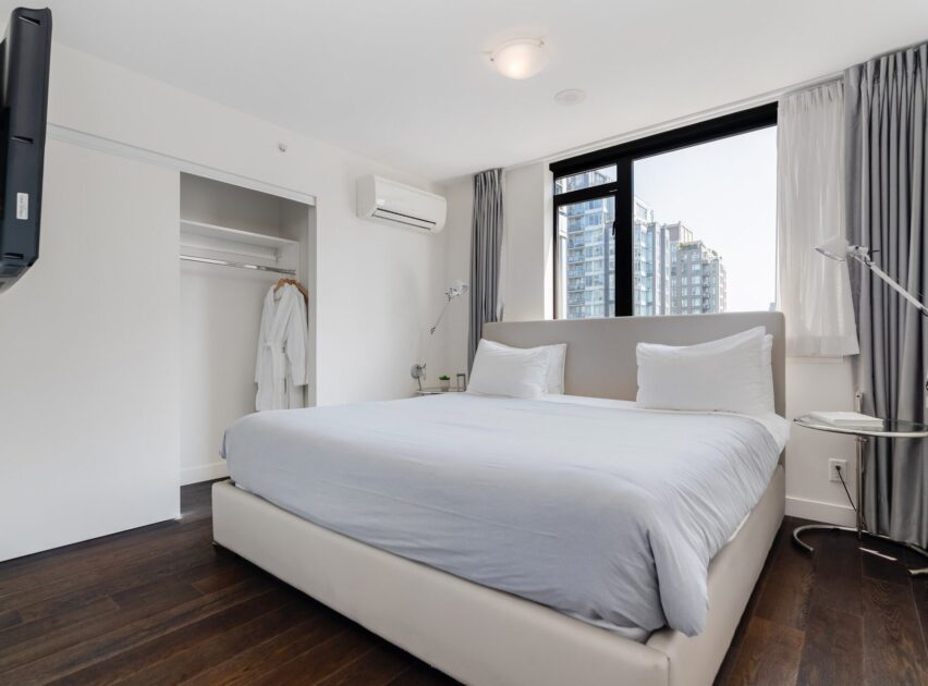 Modern bedroom with a large bed, white linens, two pillows, a wall-mounted TV, open closet with bathrobes, floor lamp, large window with city view, gray curtains, and dark wood floor.
