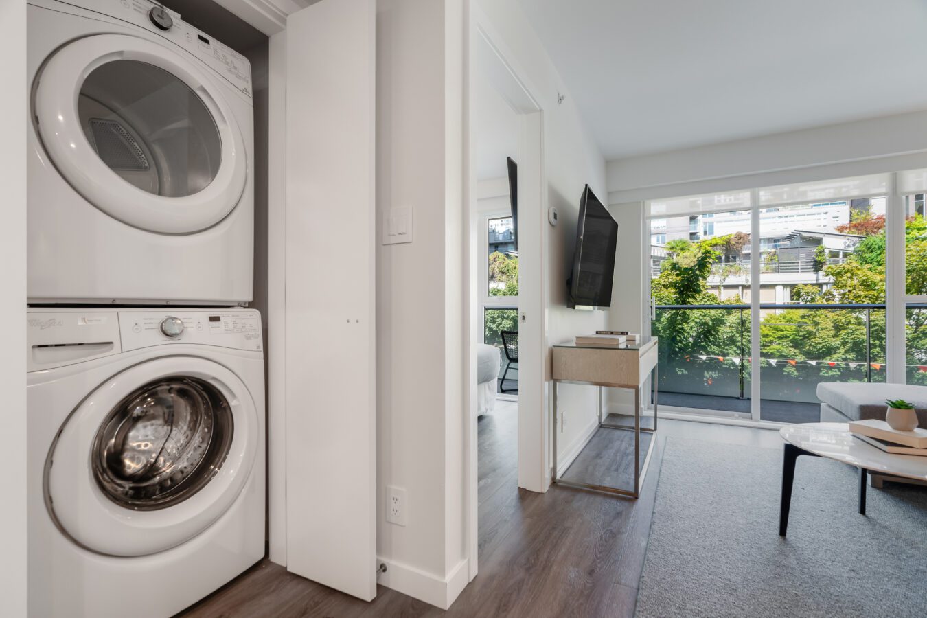A modern apartment with a stacked washer and dryer in a closet, adjoining a bright living room with large windows, a wall-mounted TV, and a view of greenery outside.