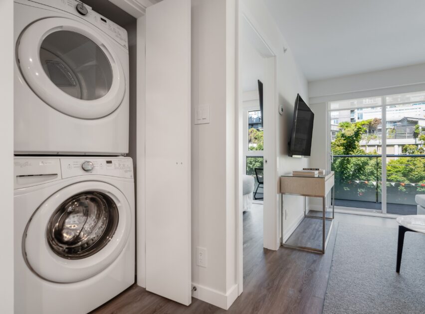 A stacked washer and dryer are built into a closet near a hallway, with a modern living area and large windows overlooking greenery in the background.