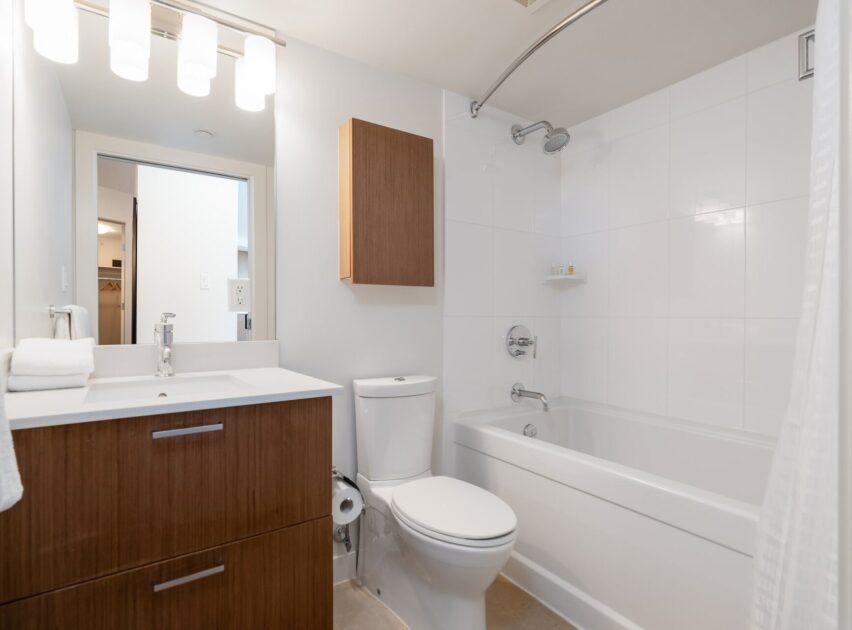 Modern bathroom with white walls and tile, a bathtub with a shower, a toilet, a wooden vanity with a sink, a wall-mounted cabinet, and a large mirror with three lights above it. Towels and toiletries are neatly arranged.