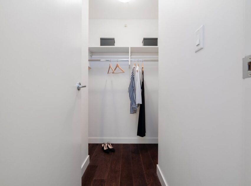 A minimal walk-in closet with white walls, dark wooden floor, a few garments and hangers on a rod, two boxes on the shelf, and a pair of black shoes on the floor.