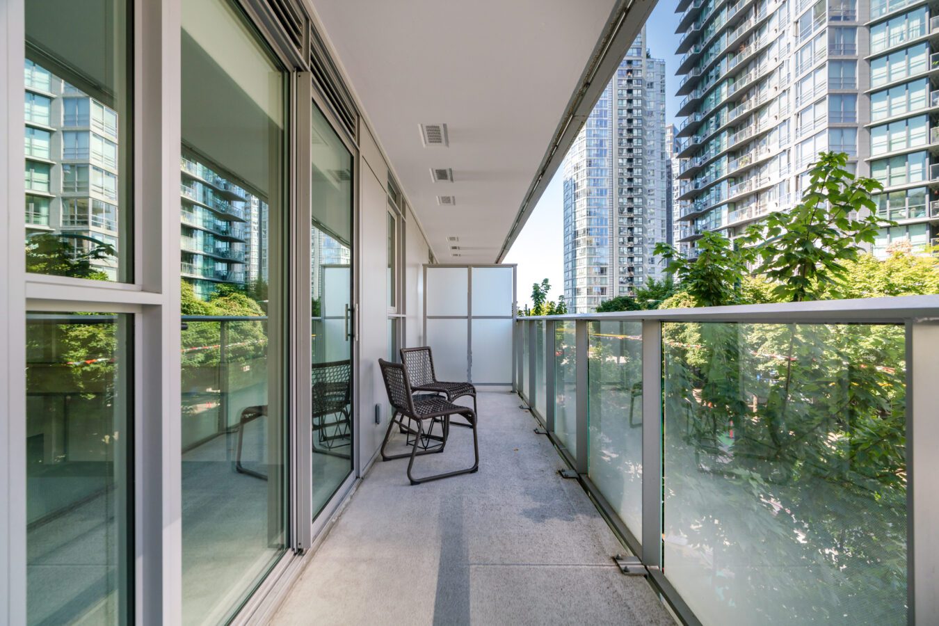 A modern apartment balcony with glass railings, two black chairs, and views of tall glass buildings and green trees in the background on a bright, sunny day.