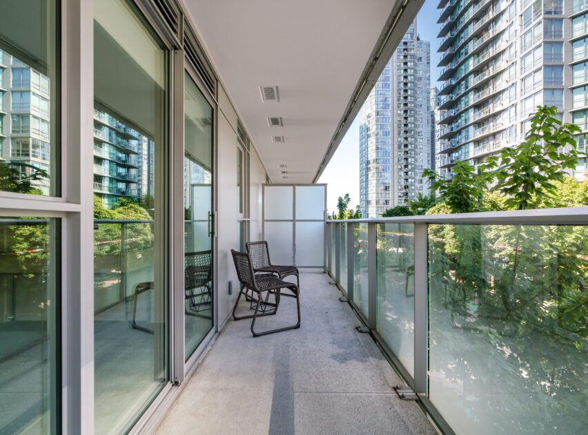 A modern apartment balcony with glass railings, two black chairs, and a view of nearby high-rise buildings and greenery on a clear day.
