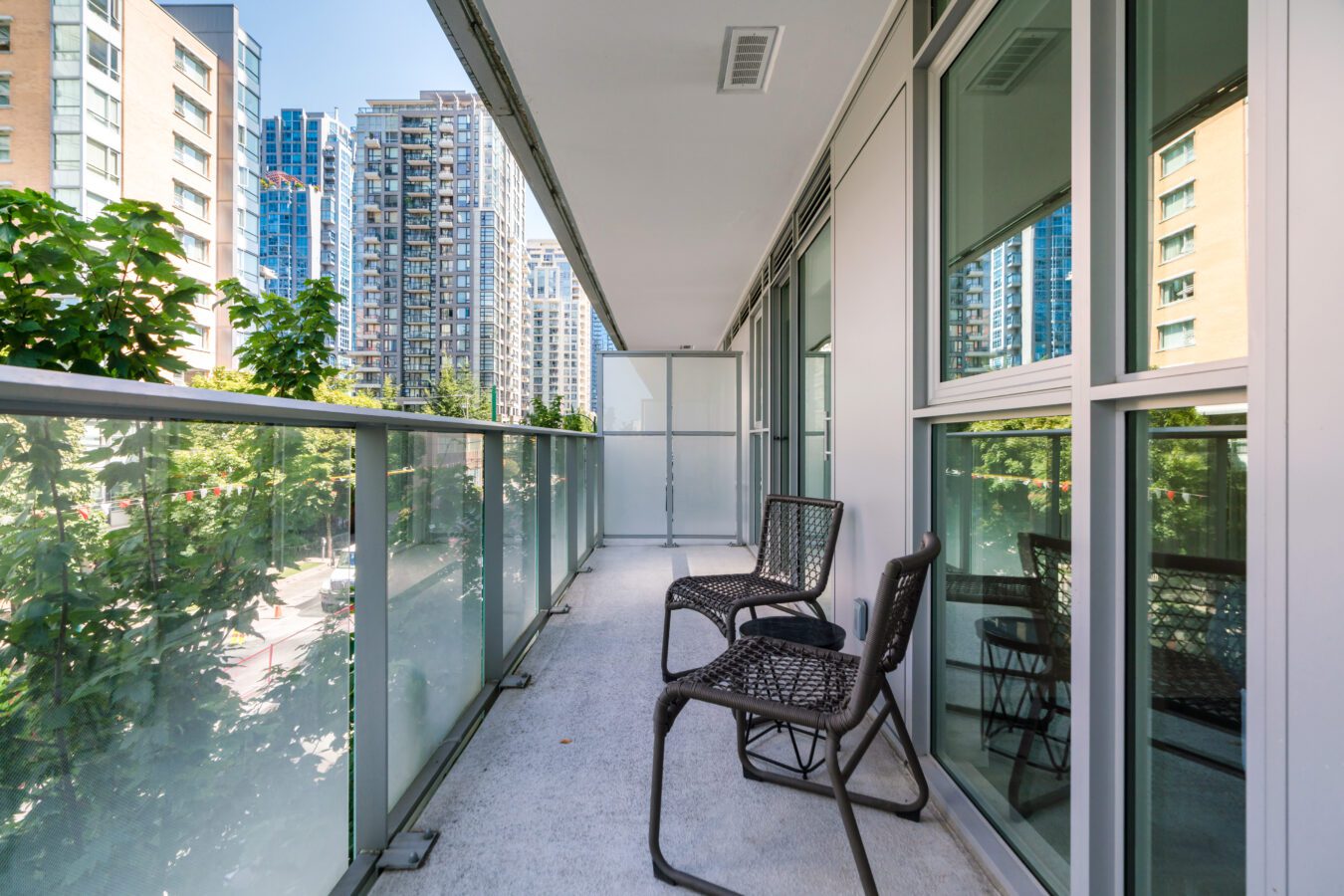 A modern apartment balcony with two black chairs, glass railing, and a view of tall city buildings and trees on a sunny day.