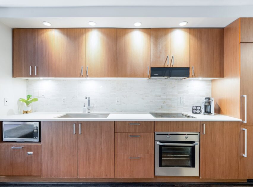 Modern kitchen with wooden cabinets, built-in oven, microwave, sink, coffee maker, and stove. White countertop and backsplash, under-cabinet lighting, and a small plant add to the clean, minimalist design.