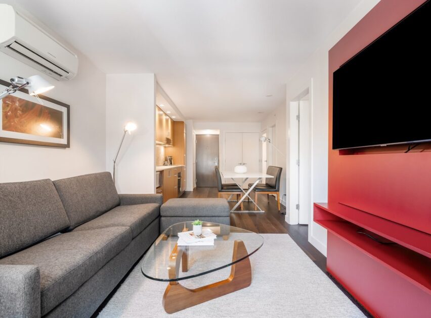 Modern living room with a gray sectional sofa, glass coffee table, wall-mounted TV on a red accent wall, and an open kitchen and dining area in the background. The space is bright and neatly organized.