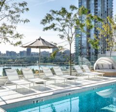 Rooftop pool with white lounge chairs, an umbrella, and a round daybed, overlooking a city skyline and river with trees and tall glass buildings in the background.