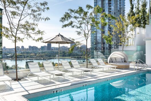 Rooftop pool with white lounge chairs, an umbrella, and a round daybed, overlooking a city skyline and river with trees and tall glass buildings in the background.