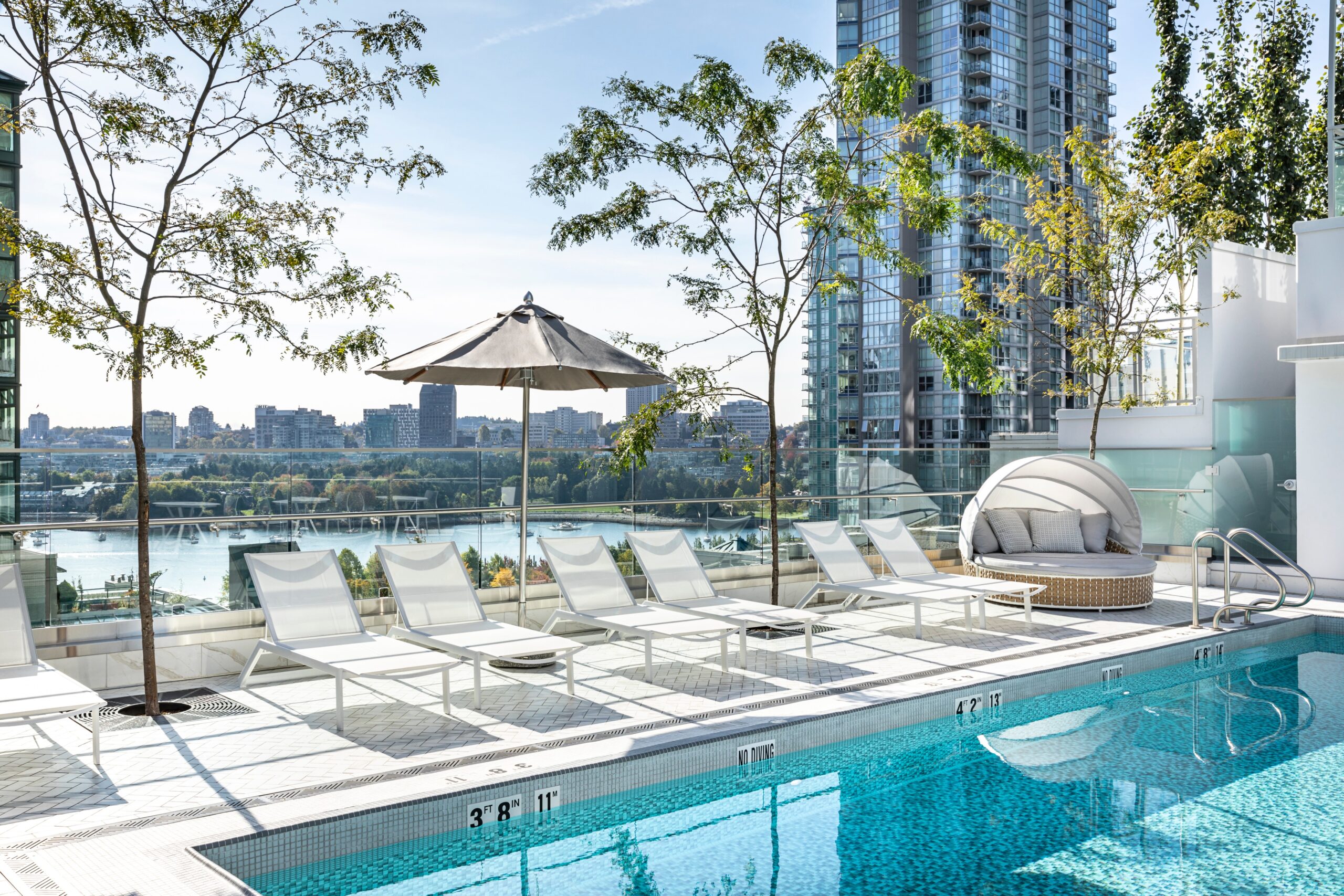 Rooftop pool with white lounge chairs, an umbrella, and a round daybed, overlooking a city skyline and river with trees and tall glass buildings in the background.
