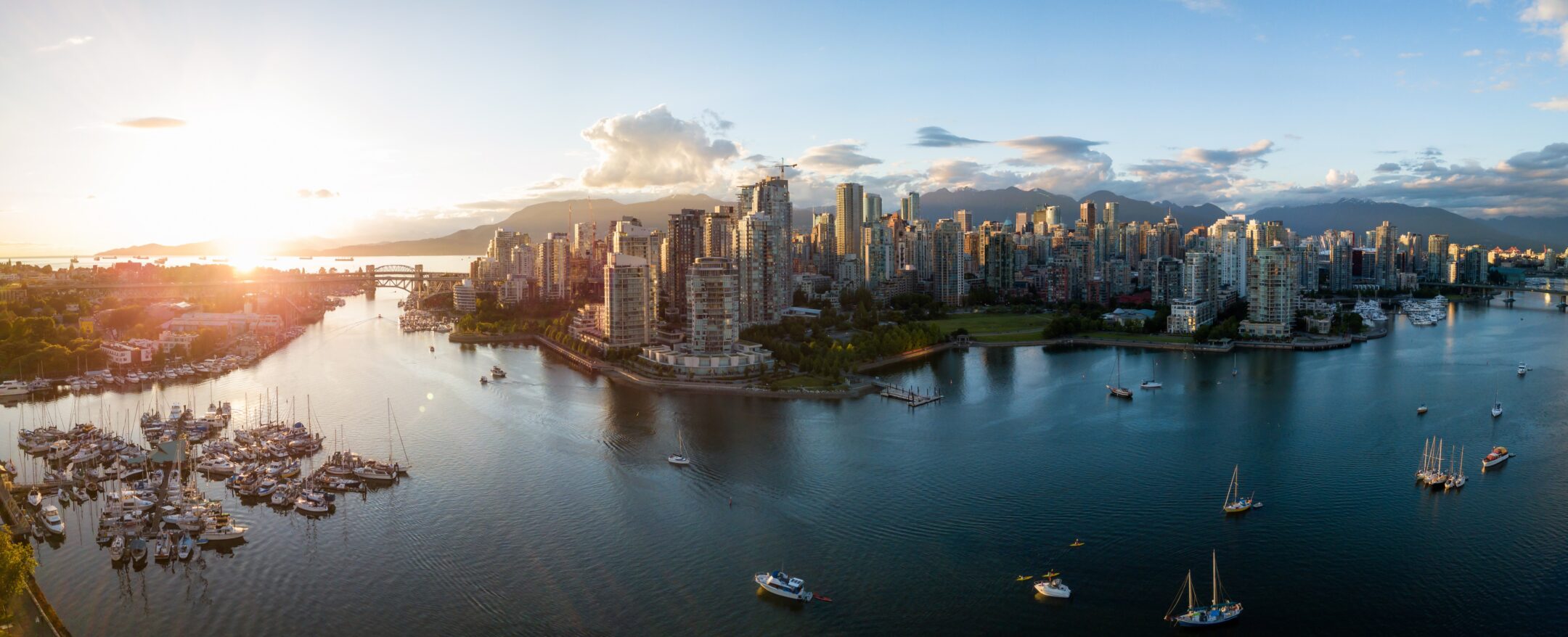Aerial view of downtown Vancouver at sunset, with tall buildings, boats in the harbor, and mountains in the background under a partly cloudy sky.