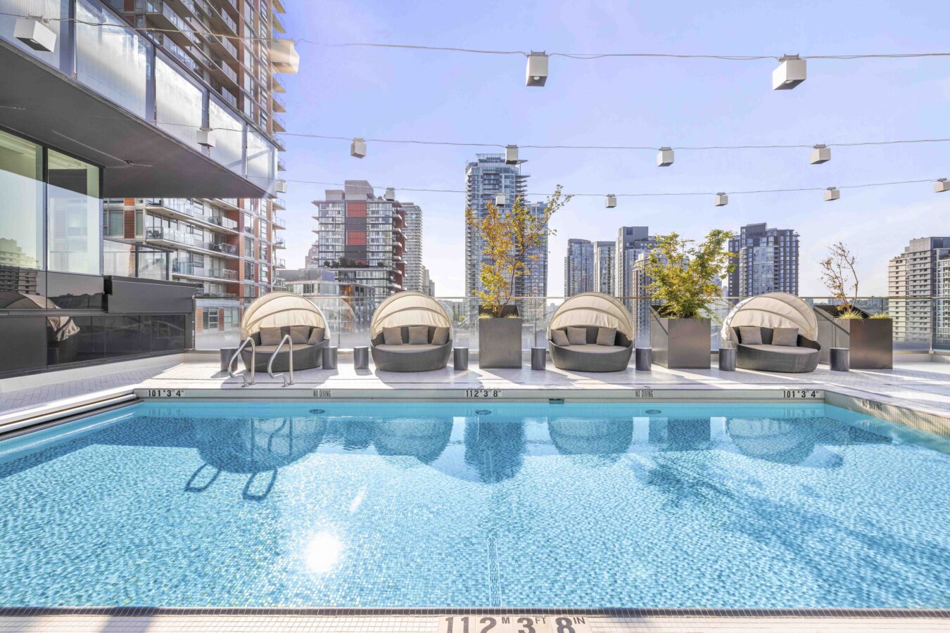 A modern rooftop pool with clear blue water, lined with four round lounge chairs and potted plants, surrounded by tall city buildings under a bright sky.