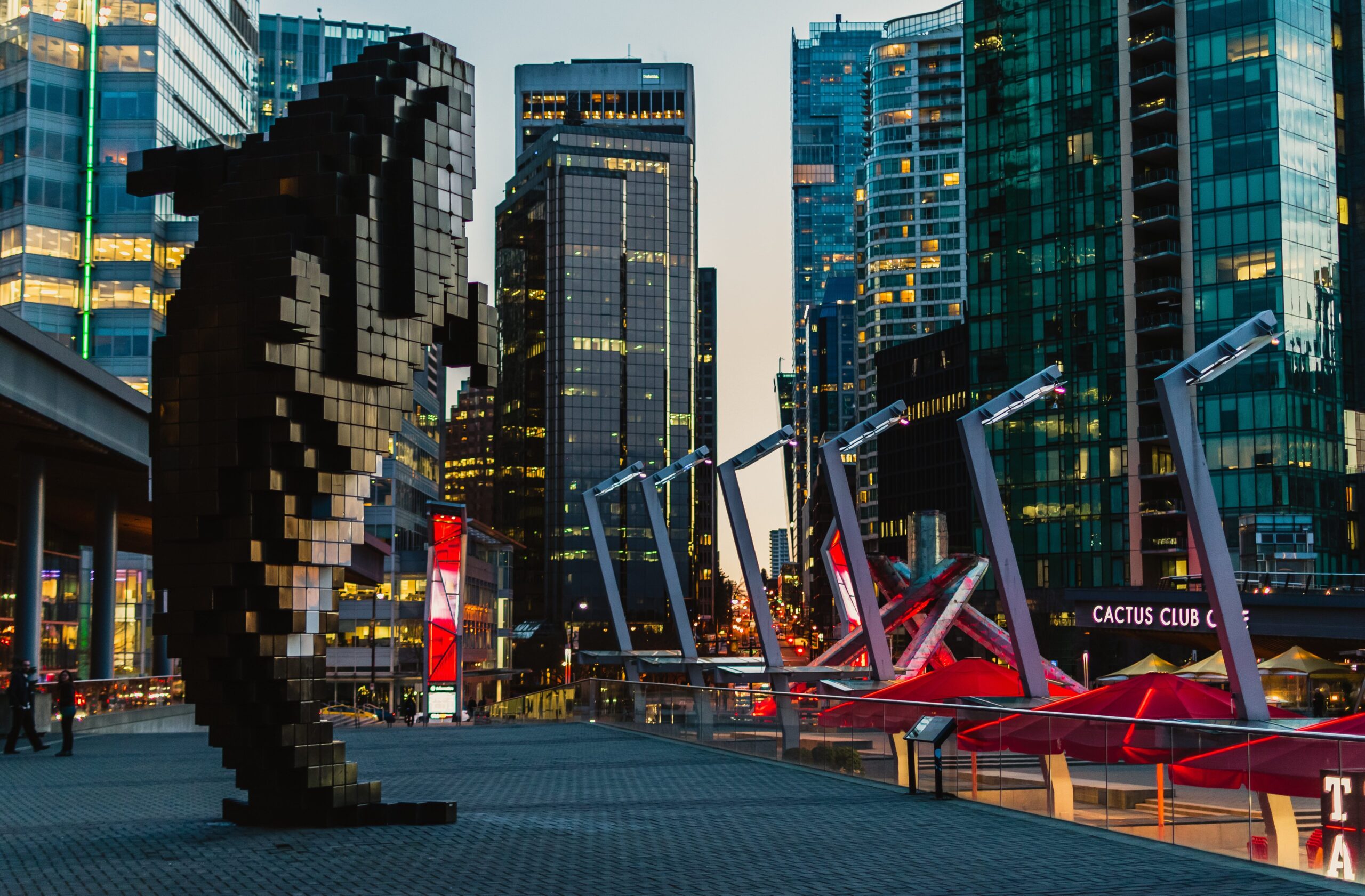 A large pixelated orca whale sculpture stands on a plaza surrounded by modern glass skyscrapers at dusk in a city. Red canopies and the Cactus Club Cafe are visible in the background.