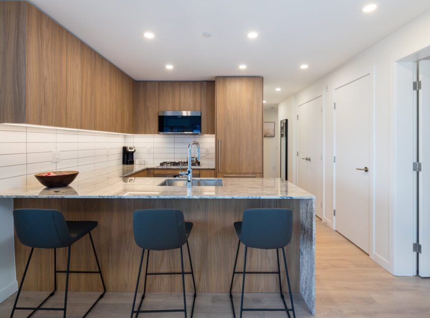 Modern kitchen with wood cabinets, white tiled backsplash, granite countertop, built-in appliances, and an island with three black barstools. The space is well-lit with recessed ceiling lights.