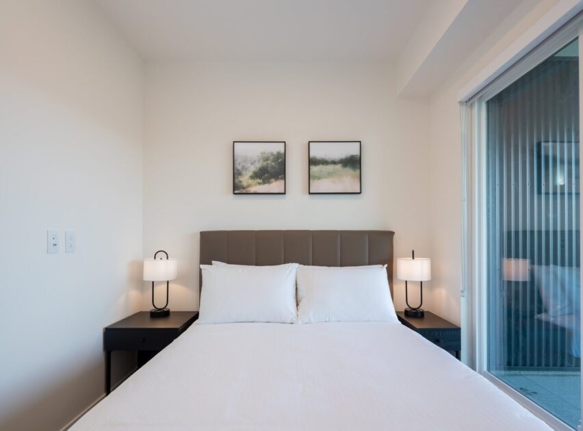 A neatly made bed with white linens is flanked by two black nightstands and matching lamps. Two abstract landscape paintings hang on the white wall above the bed. A glass door is visible to the right.